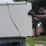 A man wearing gloves and a black shirt is seen handling refrigerator removal, pushing a large white appliance outdoors near houses and trees on a cloudy day.
