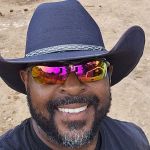 A man wearing a black cowboy hat and reflective sunglasses smiles at the camera while standing outdoors on a dirt surface, ready to assist with Arlington junk removal service.