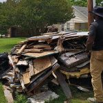 A man in a black shirt and tan pants stands beside a large pile of discarded wood, debris, and furniture from a demolition cleanup on a grassy curb in a residential neighborhood. Houses and trees are visible in the background.