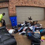 A man in a green shirt and tan pants sorts through a pile of trash, furniture, and debris on the driveway, possibly preparing for junk removal, in front of a brick house with three closed garage doors.