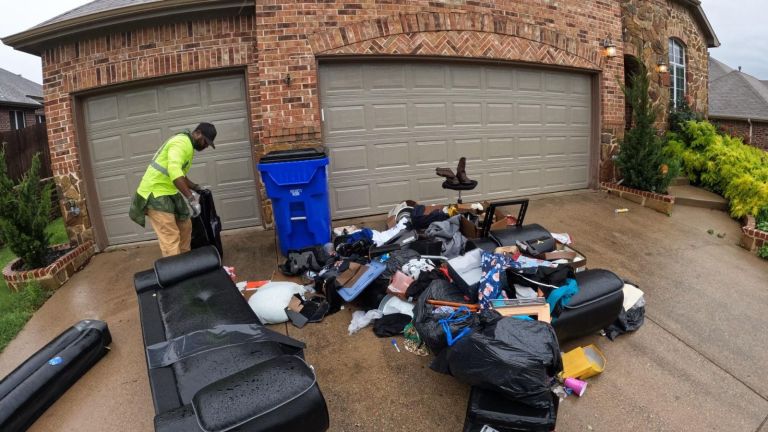 A man in a green shirt and tan pants sorts through a pile of trash, furniture, and debris on the driveway, possibly preparing for junk removal, in front of a brick house with three closed garage doors.