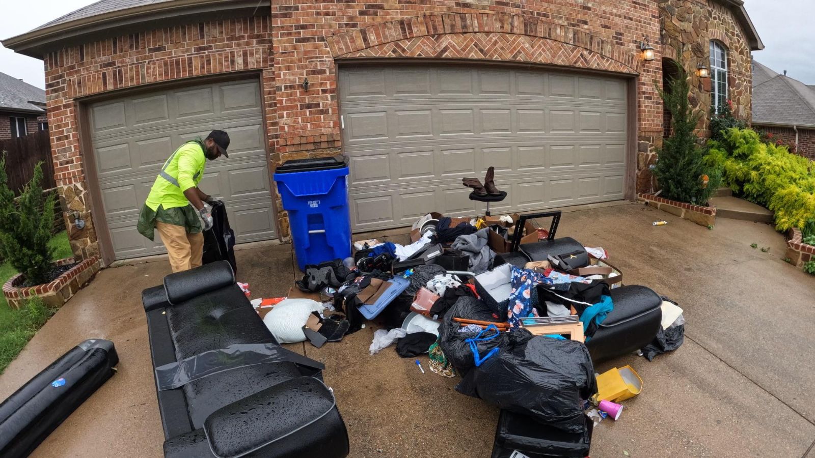 A man in a green shirt and tan pants sorts through a pile of trash, furniture, and debris on the driveway, possibly preparing for junk removal, in front of a brick house with three closed garage doors.