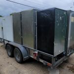 A trailer loaded with old refrigerators and freezers, ready for refrigerator removal, is parked on a street near industrial buildings, with a white truck in front and cloudy skies overhead.