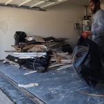 A man tackling a garage cleanout holds a large black trash bag, standing near a pile of wood scraps and debris on the garage floor as sunlight streams in through the open door.