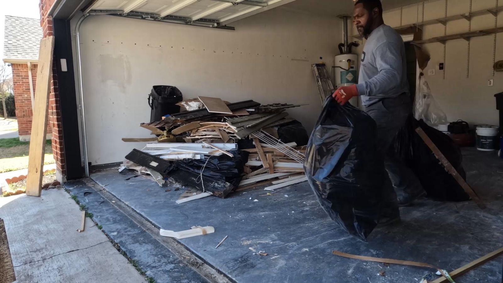 A man tackling a garage cleanout holds a large black trash bag, standing near a pile of wood scraps and debris on the garage floor as sunlight streams in through the open door.