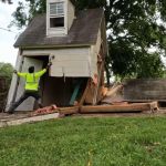 A person in a neon yellow shirt works on shed removal, pulling at the corner of a small, partially collapsed house with debris scattered on the grass and a large tree in the background.