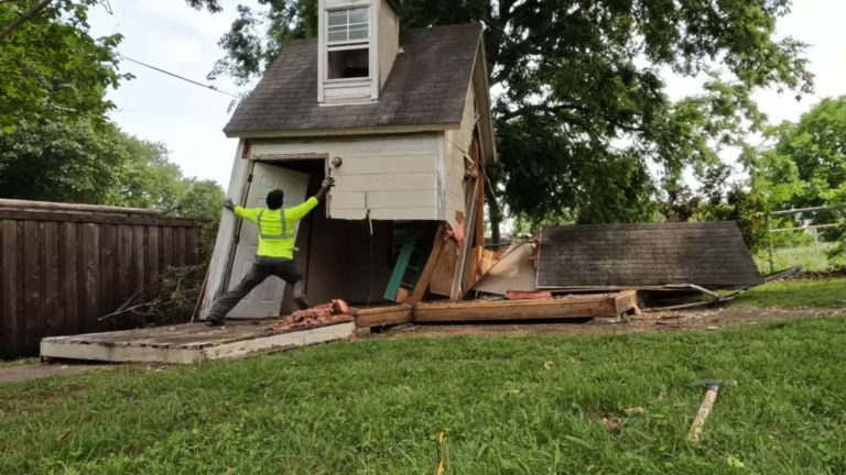 A person in a neon yellow shirt works on shed removal, pulling at the corner of a small, partially collapsed house with debris scattered on the grass and a large tree in the background.