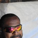 A man wearing reflective sunglasses stands in front of a brick wall, smiling, with a large, plastic-wrapped mattress behind him, ready for mattress removal.