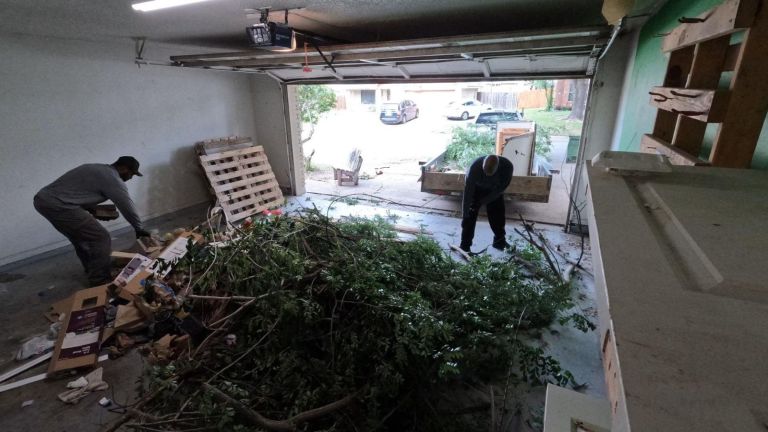 Two people tackle a garage cleanout, sorting through branches, leaves, and cardboard. With the garage door open to the driveway and trees outside, daylight brightens their organized efforts.