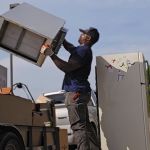 A man lifts an old refrigerator onto a trailer filled with discarded appliances and boxes, part of a junk removal service. Another fridge with magnets stands nearby, while trees are visible in the background beneath a clear sky.
