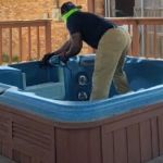 Wearing protective gear, a person uses a power tool for hot tub removal, cutting apart a blue spa on a wooden deck outside a house.
