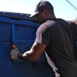 A man wearing a black shirt, gloves, and a cap is pushing a large blue dumpster outdoors on a sunny day, showcasing efficient junk removal service.