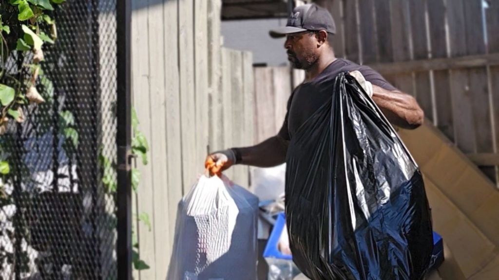 A man wearing a black cap and shirt carries two large garbage bags, one white and one black, in an outdoor area next to a wooden fence and gate—typical of a junk removal service in progress.