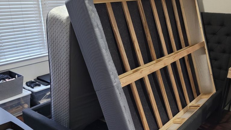 A mattress and a wooden bed frame, awaiting mattress removal, lean upright against each other in a room with a window, storage bins, and part of a gray upholstered bed visible.