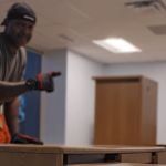 A person wearing gloves, a gray shirt, and a black head covering stands in an office with wooden furniture, possibly discussing a junk removal service. The slightly out-of-focus background shows a large window and a cabinet.