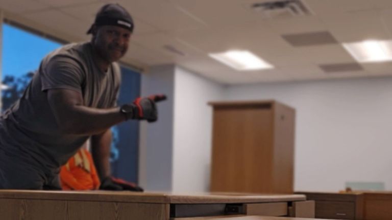 A person wearing gloves, a gray shirt, and a black head covering stands in an office with wooden furniture, possibly discussing a junk removal service. The slightly out-of-focus background shows a large window and a cabinet.