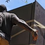 A person wearing gloves and a cap performs refrigerator removal, moving a large fridge secured with straps outdoors under a clear blue sky.