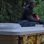 A man in black clothes and gloves uses a power tool for hot tub removal, dismantling the top of an outdoor hot tub with exposed insulation and wires inside. Green foliage is in the background.