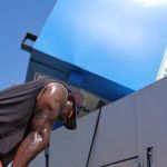 A man wearing a black sleeveless shirt and red shorts leans forward, sweating, in front of a large blue donation box removal container under a clear sky.