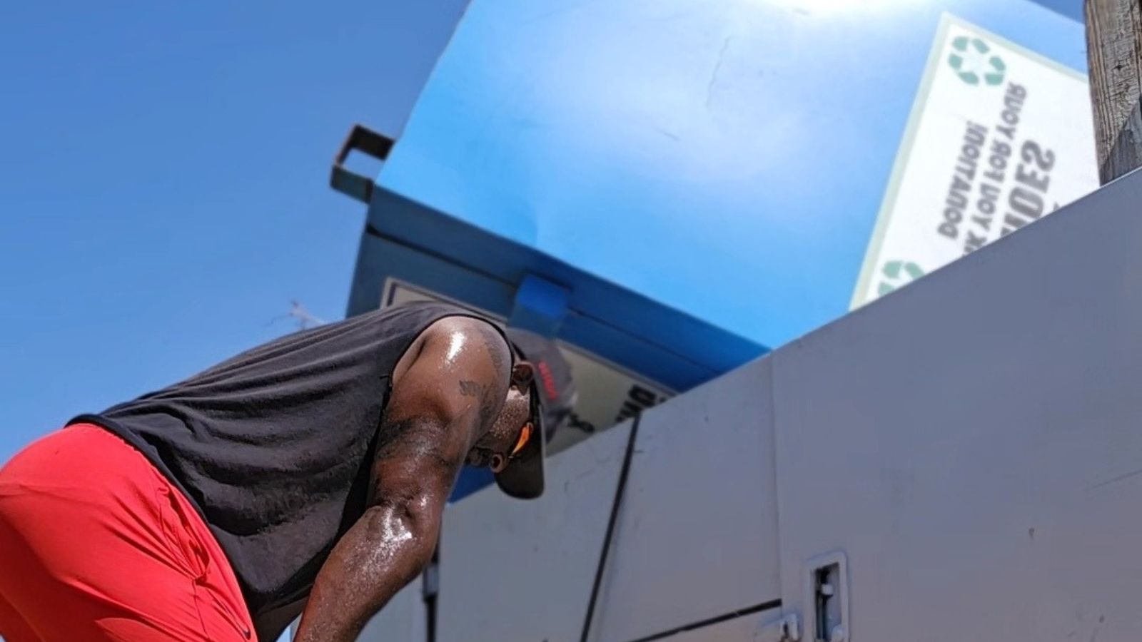 A man wearing a black sleeveless shirt and red shorts leans forward, sweating, in front of a large blue donation box removal container under a clear sky.