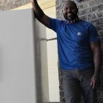 A man wearing a blue t-shirt and dark pants stands outdoors, smiling and leaning with one arm on a large white refrigerator next to a brick wall, ready for refrigerator removal.