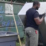 A man wearing gloves stands next to an open dumpster and a large metal object, smiling, possibly after a refrigerator removal. Nearby are a broom, a gray trash bin, and graffiti on the dumpster that reads NO SCRAPING. Green foliage is in the background.