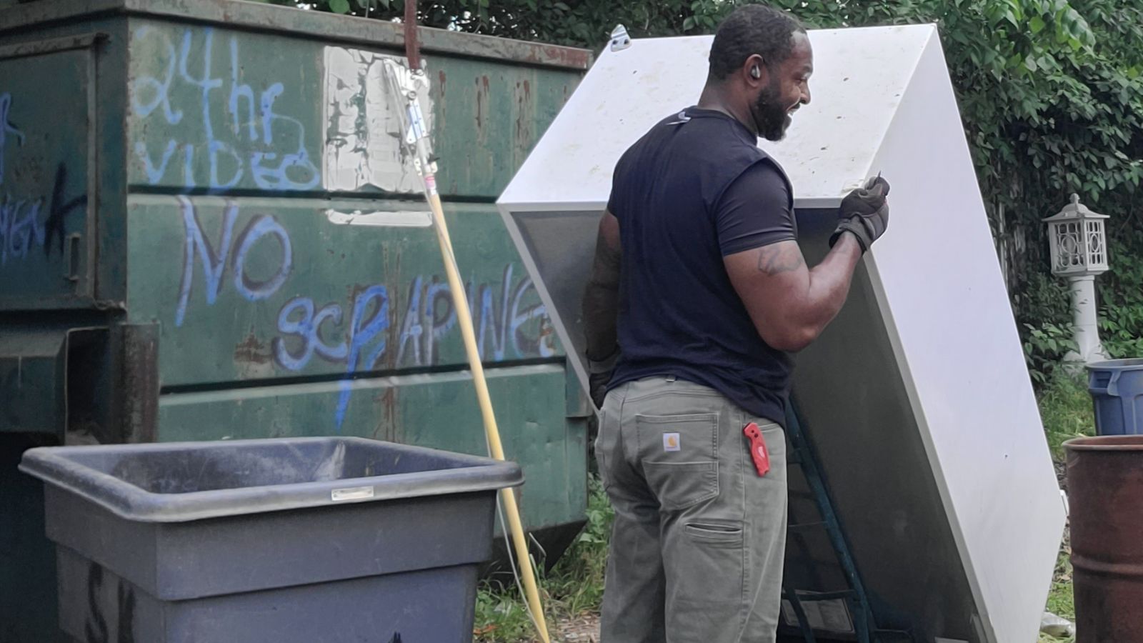A man wearing gloves stands next to an open dumpster and a large metal object, smiling, possibly after a refrigerator removal. Nearby are a broom, a gray trash bin, and graffiti on the dumpster that reads NO SCRAPING. Green foliage is in the background.