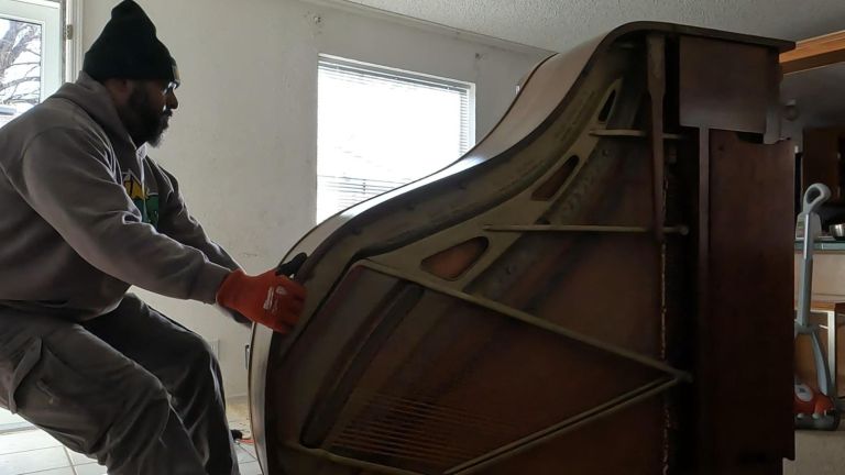 A person wearing a beanie, hoodie, and gloves carefully pushes an upright piano on a tiled floor near a large window in a softly lit room, demonstrating the process of piano removal.