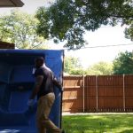 A person wearing gloves is handling a large blue hot tub on its side during a hot tub removal across a grassy backyard, with a wooden fence and trees in the background.