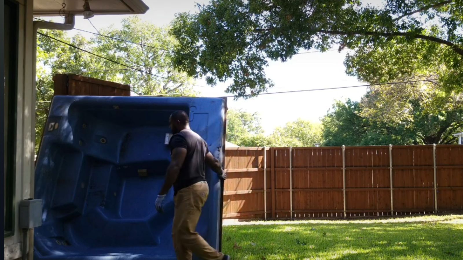 A person wearing gloves is handling a large blue hot tub on its side during a hot tub removal across a grassy backyard, with a wooden fence and trees in the background.
