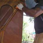 A person wearing gloves and a dark shirt handles an upright piano outside on a porch during a piano removal. Sunlight filters through the trees in the background.