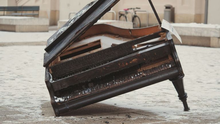 A broken grand piano with a missing leg and damaged keys sits outdoors on a snowy, paved surface; its lid is partially open, revealing the interior. A bicycle and benches are visible in the background.