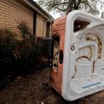 A large, white and brown hot tub lies tipped on its side outside near a brick house, surrounded by bushes and trees—possibly awaiting hot tub removal. A person in light clothing stands partially visible on the right.