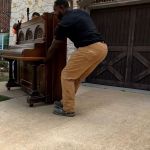 Two people are performing a piano removal on a driveway in front of a house with brick and stone exterior and brown garage doors. One person lifts while the other carefully guides the upright piano.