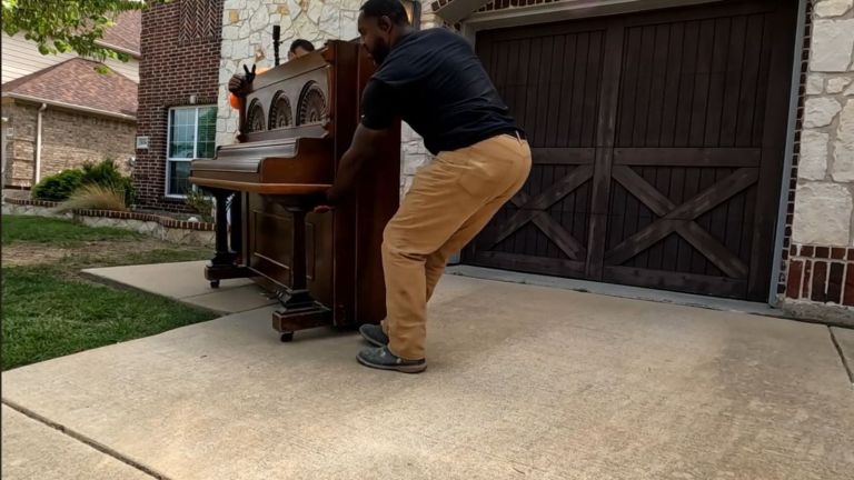 Two people are performing a piano removal on a driveway in front of a house with brick and stone exterior and brown garage doors. One person lifts while the other carefully guides the upright piano.