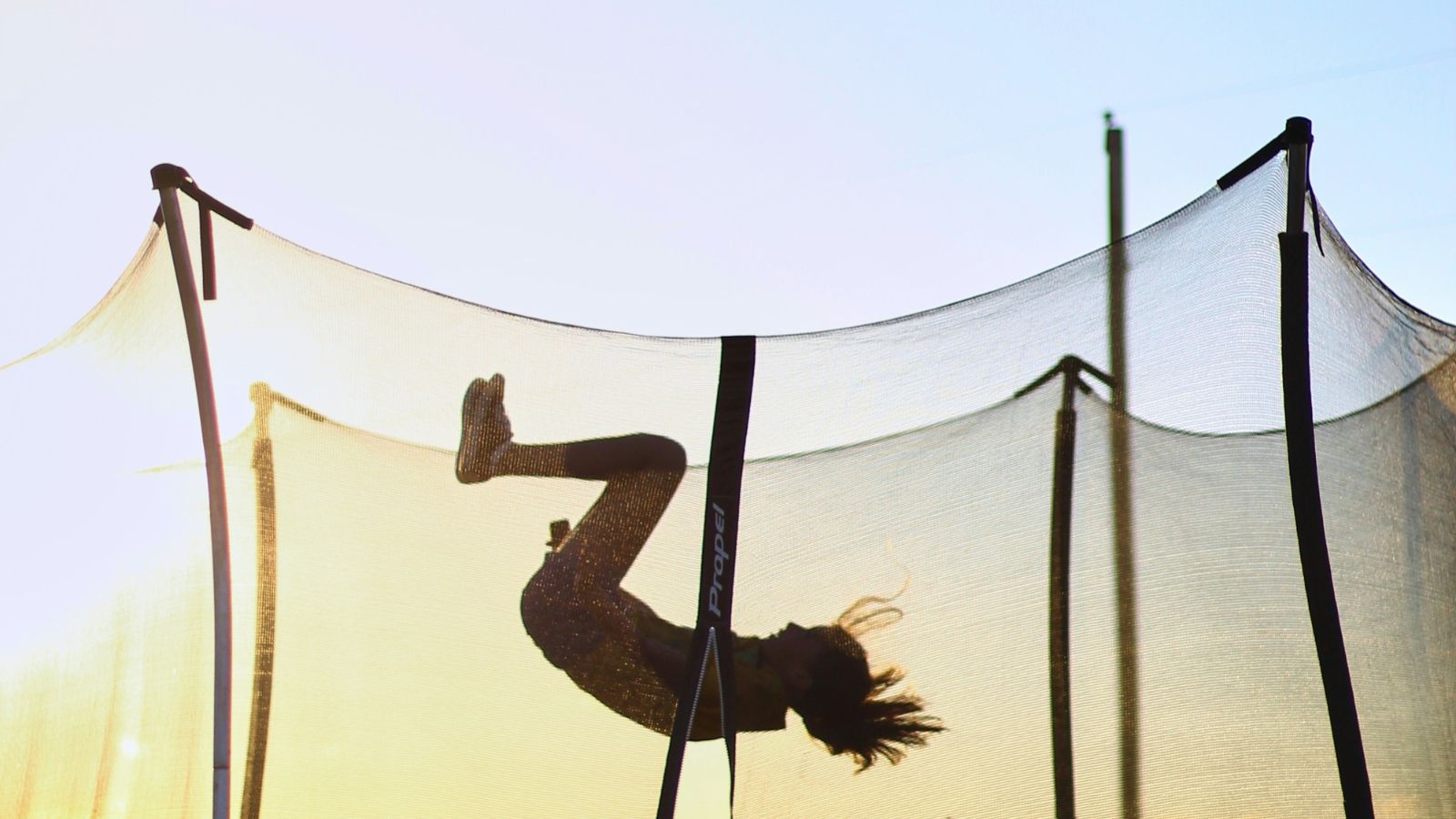 A child is captured mid-flip on a trampoline, silhouetted against the bright sky. The safety netting around the trampoline is visible, and the sunlight creates a warm, glowing background.
