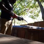A person uses a power saw to cut into the edge of a wooden hot tub outside, sunlight filtering through trees and a wooden fence in the background during a hot tub removal project.