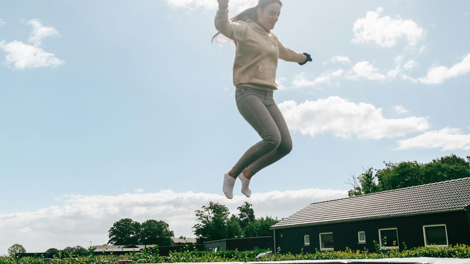 A young woman wearing a beige hoodie and gray pants jumps high on a trampoline outdoors, with houses, trees, and a partly cloudy sky in the background.