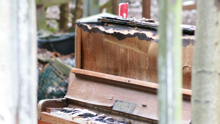 A weathered, partially burnt upright piano sits outdoors among trees, with damaged keys and a melted red candle on top. The wood is charred, and debris is scattered around, suggesting long-term exposure to the elements.