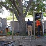 Two people stand in a backyard near a large tree; one person organizes items in a tall shelving unit while the other observes. Wooden posts, scattered materials, and signs of hot tub removal indicate construction or renovation work in progress.