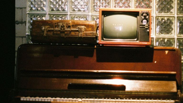 A vintage brown suitcase and a red retro television sit on top of a closed upright piano, with a glass block wall in the background.