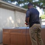 A person wearing a dark t-shirt and khaki pants stands outside next to a large covered hot tub, possibly preparing for hot tub removal, holding a cable or cord, with trees and a house in the background.