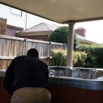 A person in a black shirt and khaki pants works on hot tub removal under a covered patio, while another stands in the background near a wooden fence and bushes.