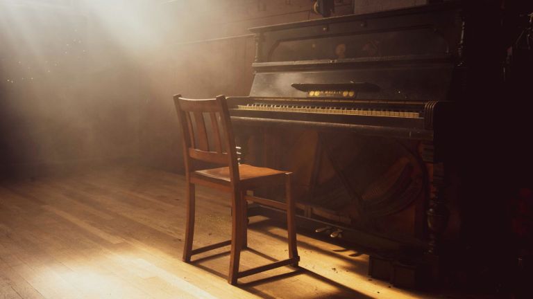 A wooden chair sits in front of an old upright piano, bathed in soft, golden sunlight streaming through a window, casting gentle shadows across a wooden floor in an empty, dusty room.