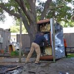 A person pushes an overturned hot tub in a backyard, tackling a hot tub removal project amid scattered tools and debris. A large tree and wooden fence enclose the area, while another person stands partially hidden behind the hot tub.