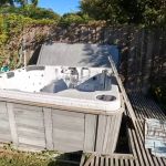 A large, empty hot tub sits in a backyard against a wooden fence, surrounded by overgrown plants and a partially collapsed slatted wooden cover—highlighting the need for hot tub removal.