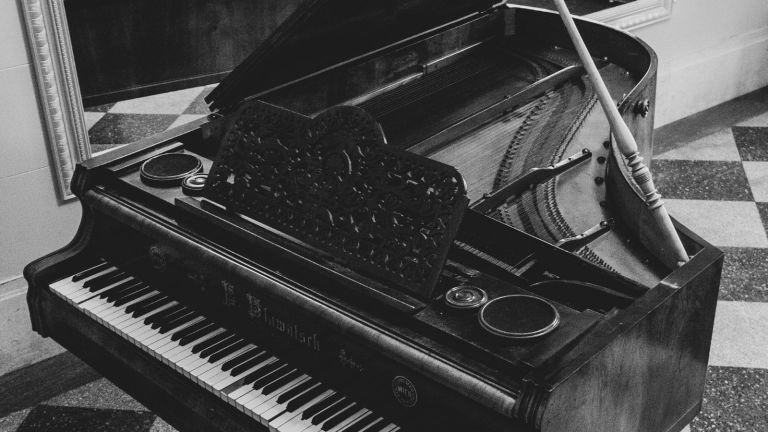 Black and white photo of a grand piano with its lid open, showing the strings and mechanisms inside, placed on a patterned floor near a wall with a framed mirror.