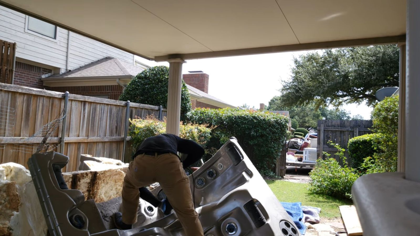 A person in tan pants and a black shirt bends over while working on hot tub removal in a backyard. The large, tilted hot tub sits under the shade of a patio cover, surrounded by a wooden fence, bushes, and a nearby house.