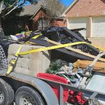 A trailer parked on a suburban street is loaded with various household items, cardboard boxes, furniture, and black garbage bags—evidence of a recent junk removal—secured with yellow straps in front of brick houses with garages.
