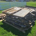A large pile of wooden boards and planks, likely from a fence removal, lies on green grass near a white picket fence, with a trailer and vehicle partially visible on the right side.